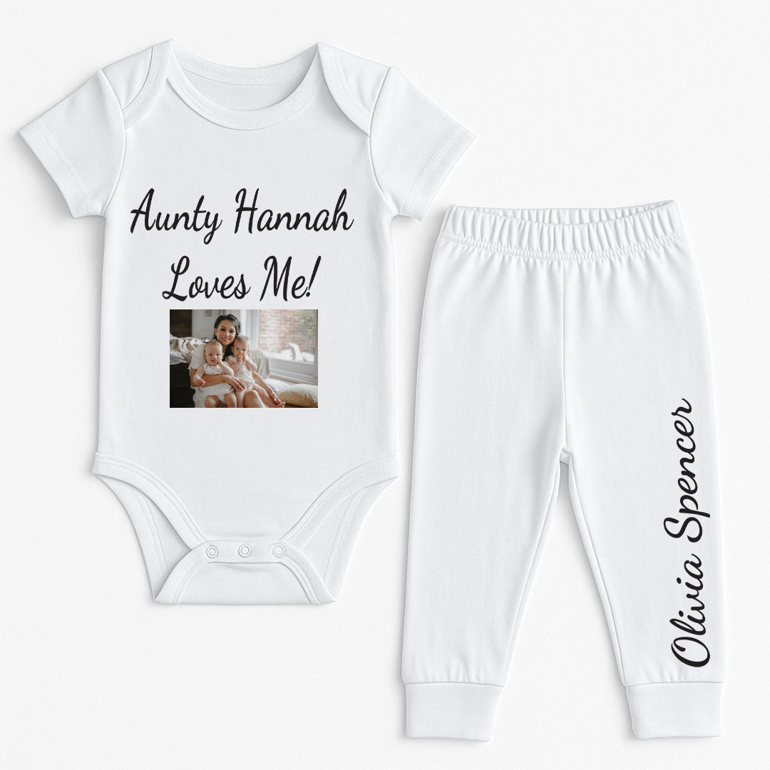 A white baby bodysuit with black text saying "Aunty Hannah Loves Me," adorned with a printed photo of a woman and a baby sitting together indoors. The bodysuit showcases vibrant, personalized prints, ideal for family gatherings and special occasions. High-quality cotton fabric enhances comfort for infants.