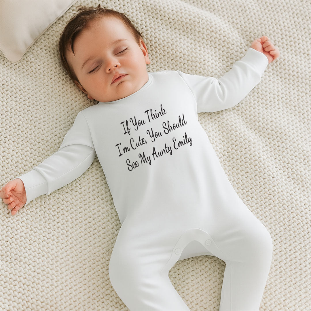 A sleeping baby lying on a textured cream knit blanket, wearing a white long-sleeve footed sleeper with the phrase 'If You Think I'm Cute, You Should See My Auntie Emily' printed in black script across the chest.