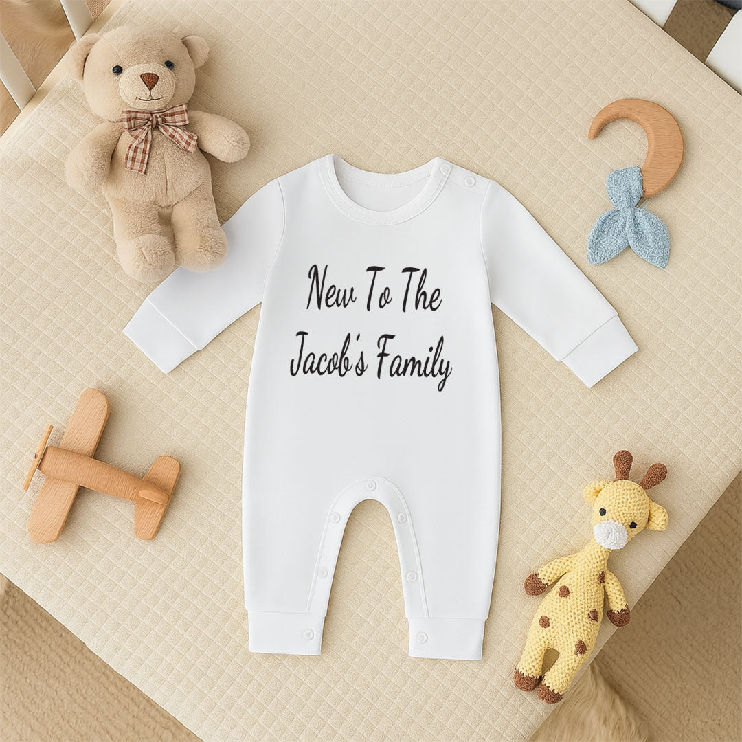 A white long-sleeve baby jumpsuit with 'New To The Jacobi's Family' printed in black script across the chest, styled in a flat lay on a beige quilted blanket with baby toys including a teddy bear, wooden airplane, crochet giraffe, and crescent moon rattle.