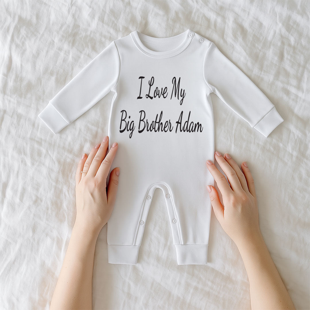 A white long-sleeve baby bodysuit with the phrase 'I Love My Big Brother Adam' printed in black script across the chest, laid flat on a white textured blanket with hands holding the sides of the garment.