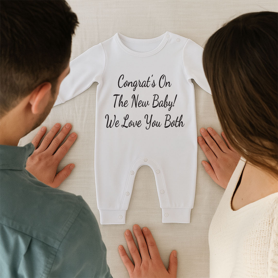 A white long-sleeve baby bodysuit with black text reading 'Congrats On The New Baby! We Love You Both' is laid flat on a light surface, with a man and woman standing on either side and touching the garment. The product is the baby bodysuit, but because a person and real environment are present, this is classified as Lifestyle.