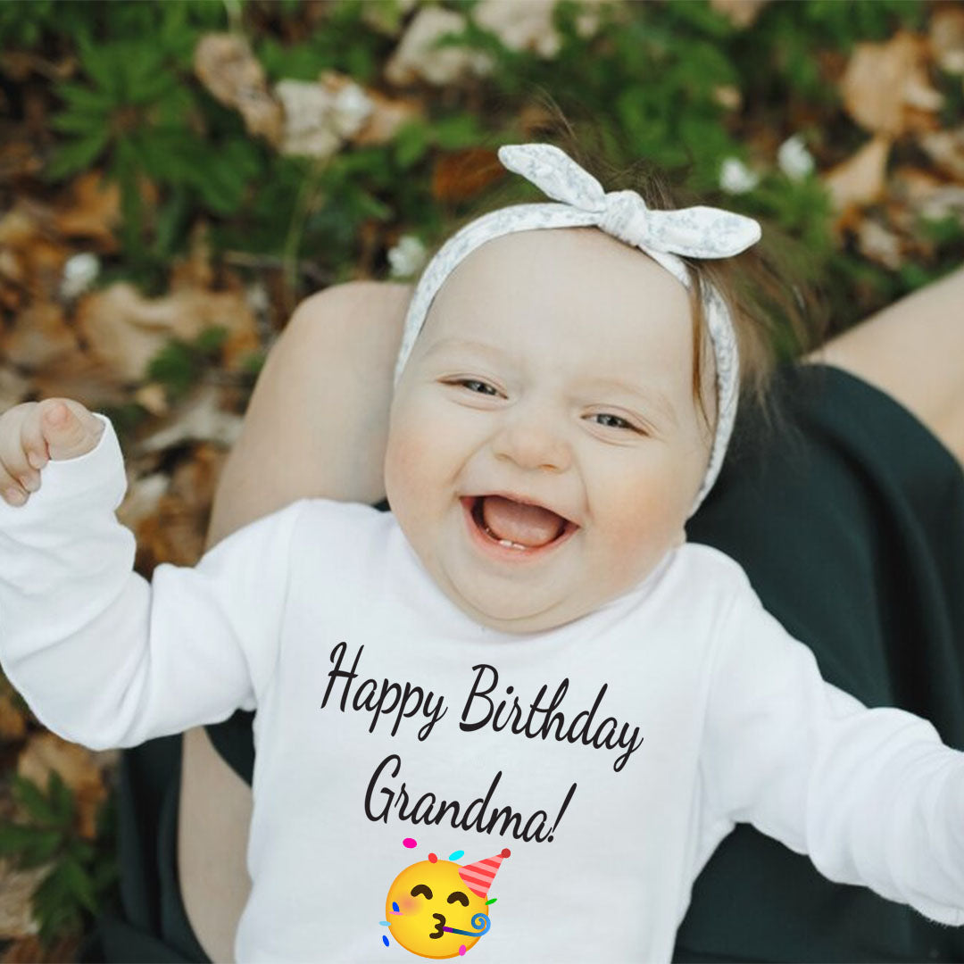 White baby onesie with joyful message “Happy Birthday Grandma!” and party face emoji, worn by a smiling baby girl outdoors.