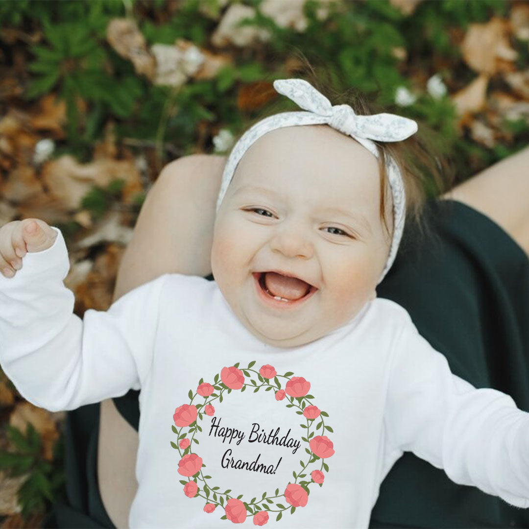 Smiling baby girl wearing a white long-sleeved bodysuit with a pink rose floral wreath and the phrase "Happy Birthday Grandma!" printed on the front.