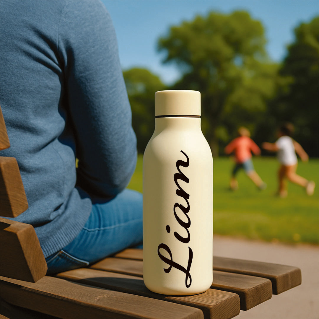 Person sitting on a wooden bench with a cream-colored water bottle labeled 'Liam' in a park setting.