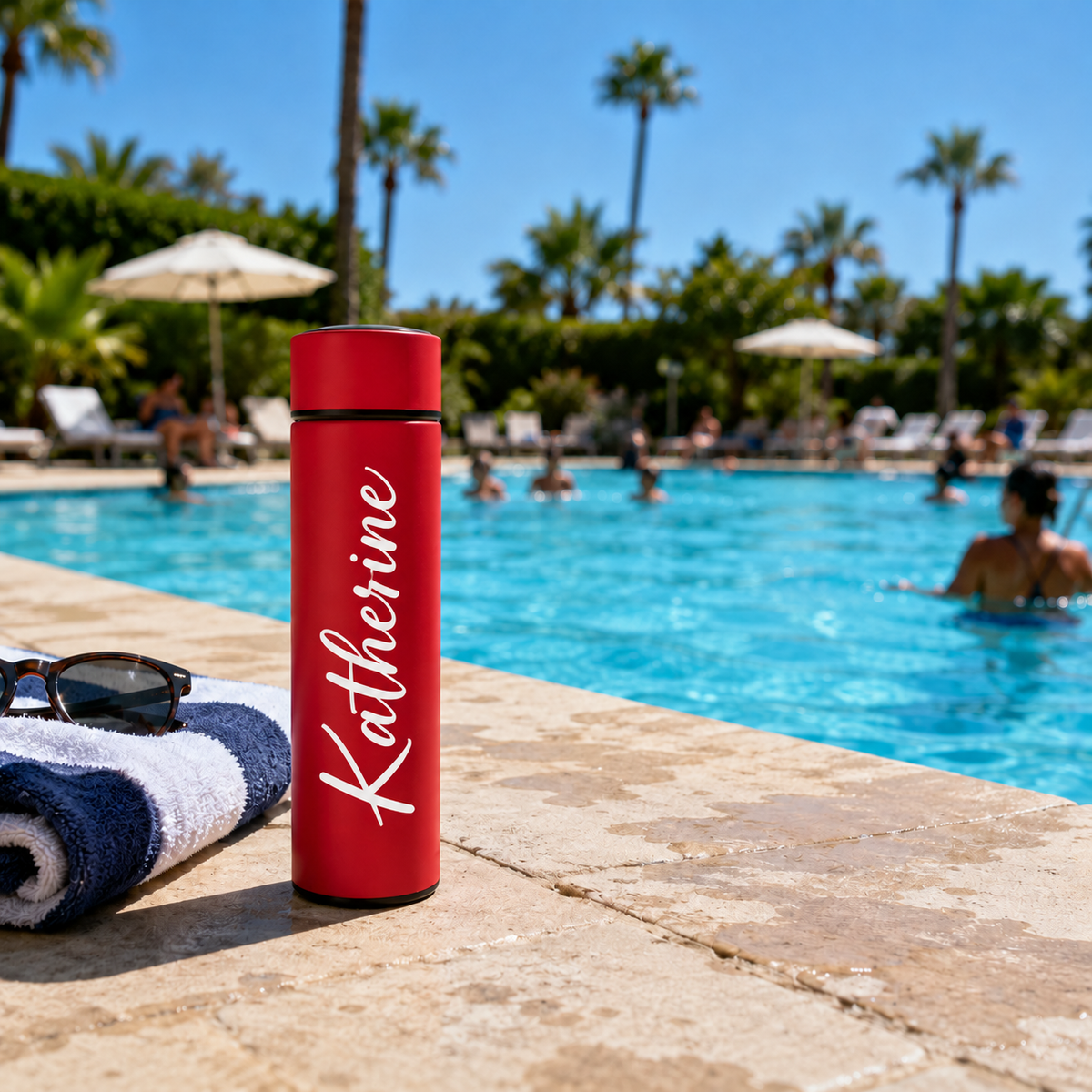 A red insulated tumbler with the name 'Katherine' printed in white script stands on a poolside deck. A rolled towel, sunglasses, and a swimming pool with palm trees are visible in the background.