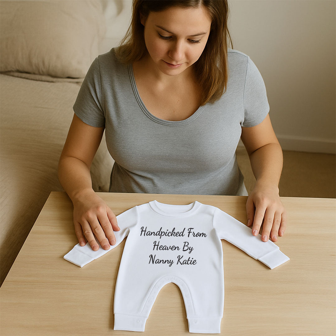 A woman sits at a light wood table, looking down at a white long-sleeve baby bodysuit laid flat. The bodysuit has black text that reads 'Handpicked From Heaven By Nanny Katie'.
