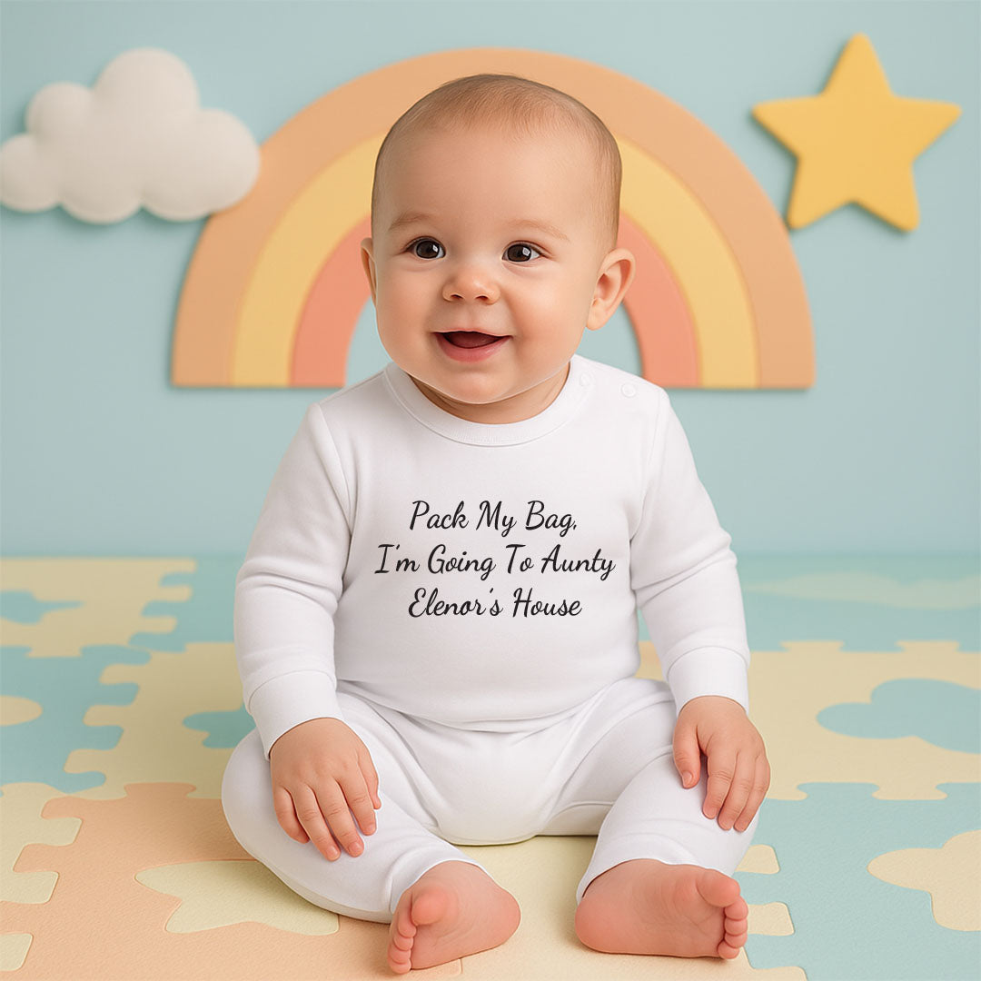 A baby sitting on pastel play mats wearing a white long-sleeve bodysuit with the text 'Pack My Bag I'm Going To Auntie Elenor's House' printed in black script. The background features a pastel blue wall with a paper rainbow, a yellow star, and a white cloud.