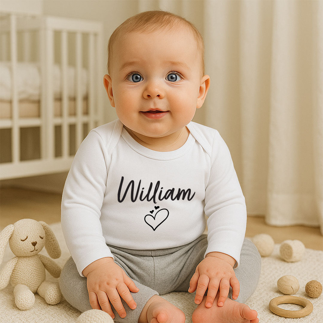 A baby sitting on a cream rug in a nursery, wearing a white long-sleeve bodysuit with the name 'William' and a small heart printed in black on the chest, paired with light gray pants.