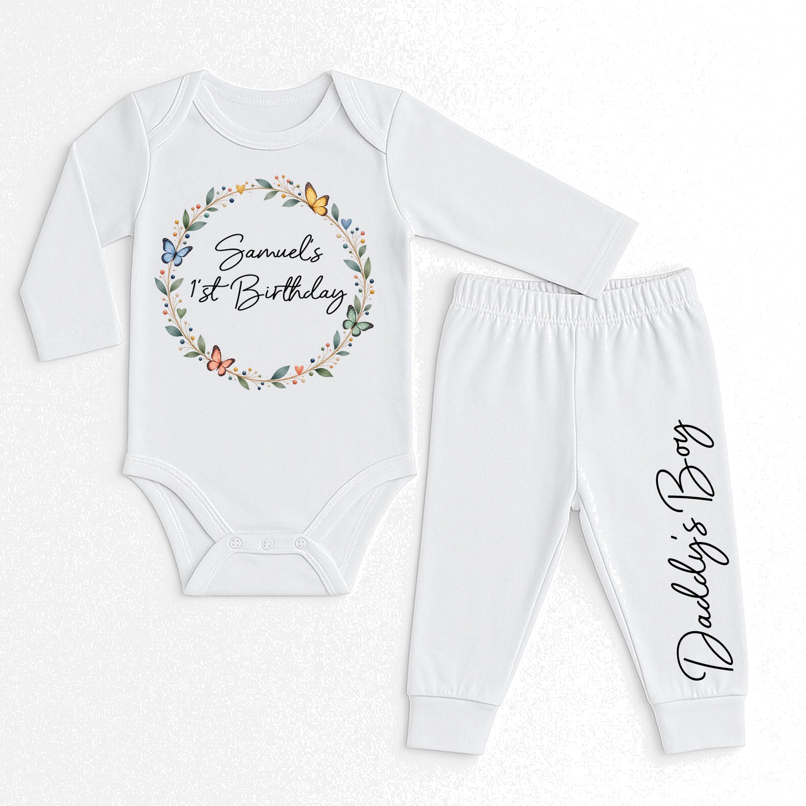 A two-piece baby outfit set consisting of a white long-sleeve bodysuit and matching white pants, laid flat on a light textured background. The bodysuit features a personalized floral wreath graphic with butterflies and the text 'Samuel's 1st Birthday'.