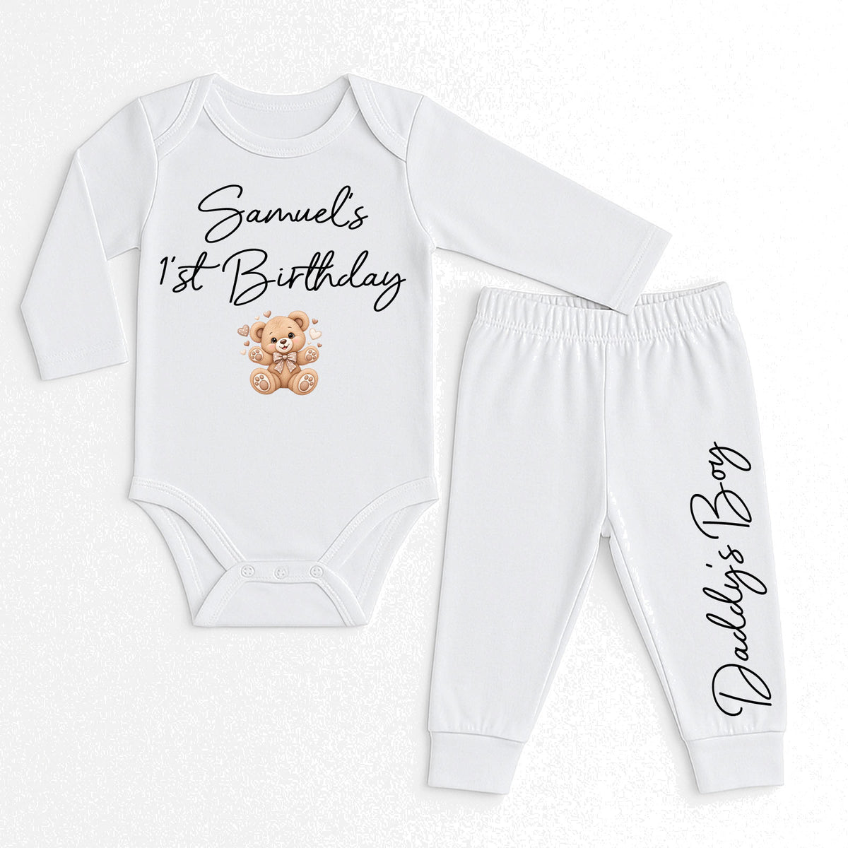 A two-piece baby outfit set consisting of a long-sleeve white bodysuit and matching jogger pants. The bodysuit features the text 'Samuel's 1st Birthday' in black script above a small brown teddy bear graphic.