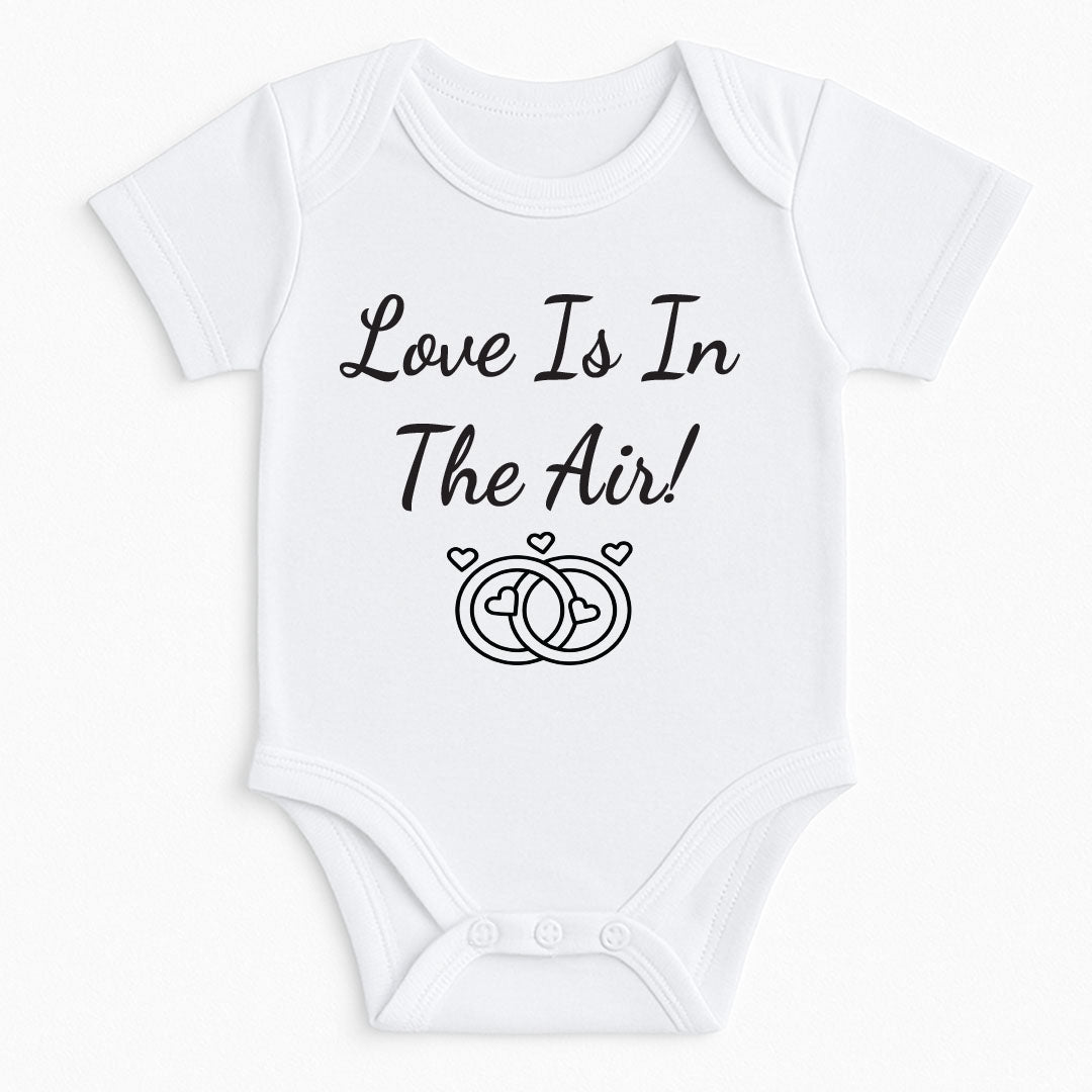 White baby bodysuit with the phrase “Love Is In The Air!” and two interlocking wedding rings surrounded by hearts.