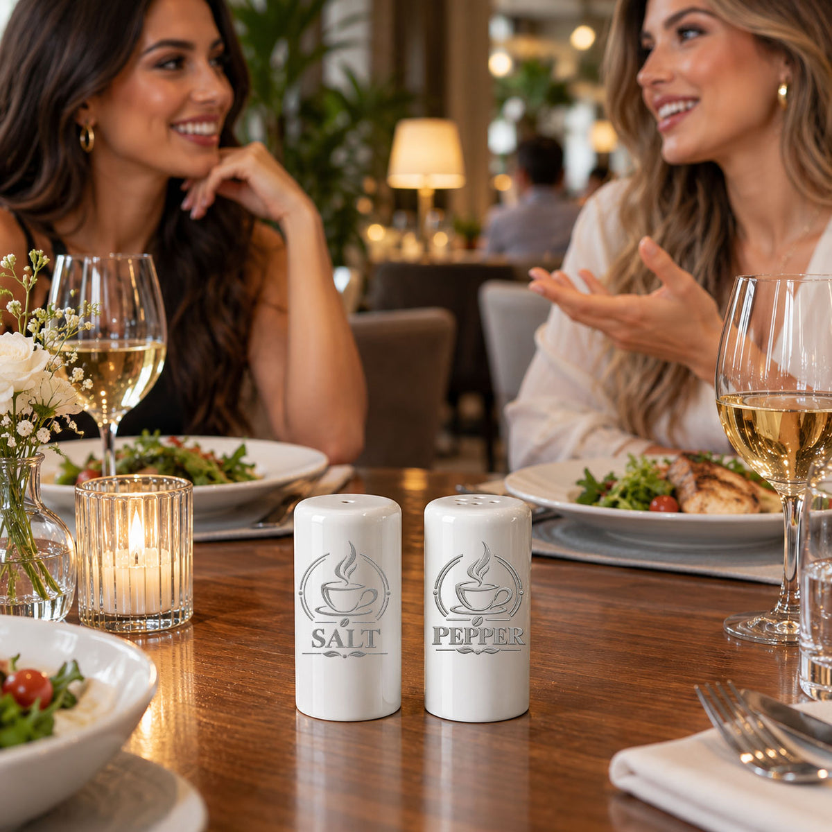 Two white salt and pepper shakers with engraved designs of a coffee cup and flame, labeled 'SALT' and 'PEPPER', on a wooden dining table in a restaurant setting with two women in the background.