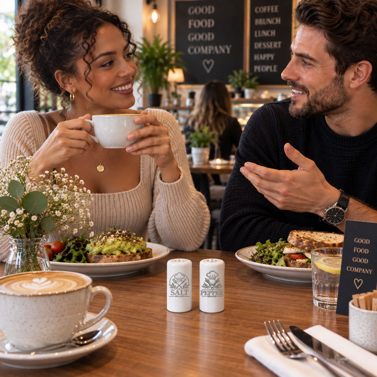 A smiling woman in a beige sweater holds a white coffee cup while sitting at a wooden cafe table with a man in a dark sweater. Plates with avocado toast and a sandwich, salt and pepper shakers, a latte, and a small sign reading GOOD FOOD GOOD COMPANY are visible.