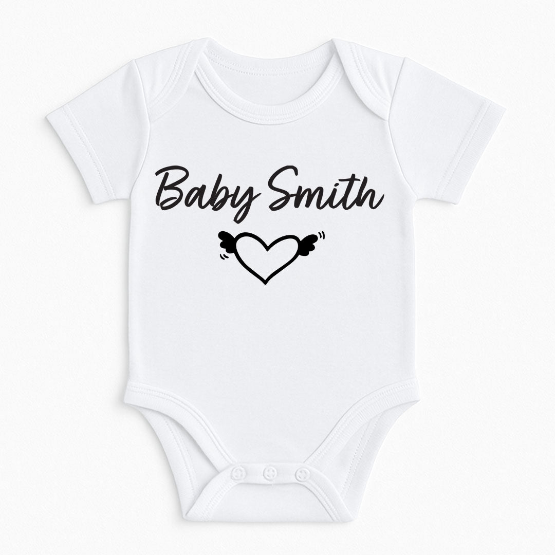 A white short-sleeve baby bodysuit with 'Baby Smith' printed in black script above a black outlined heart with small wings, shown on a plain white background.