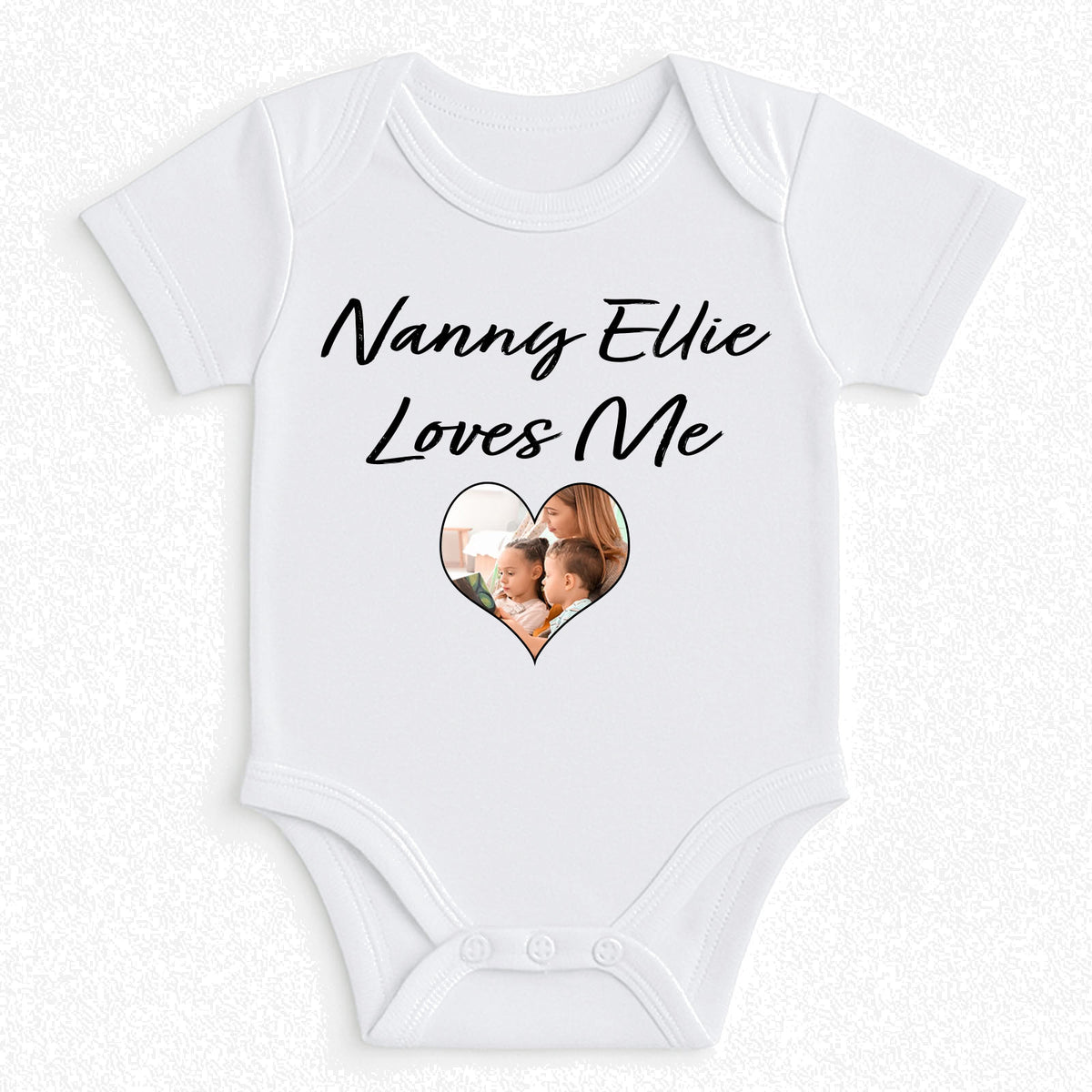 A white short-sleeve baby bodysuit with 'Nanny Ellie Loves Me' printed in black script above a heart-shaped photo window on the front, displayed flat against a light textured background.