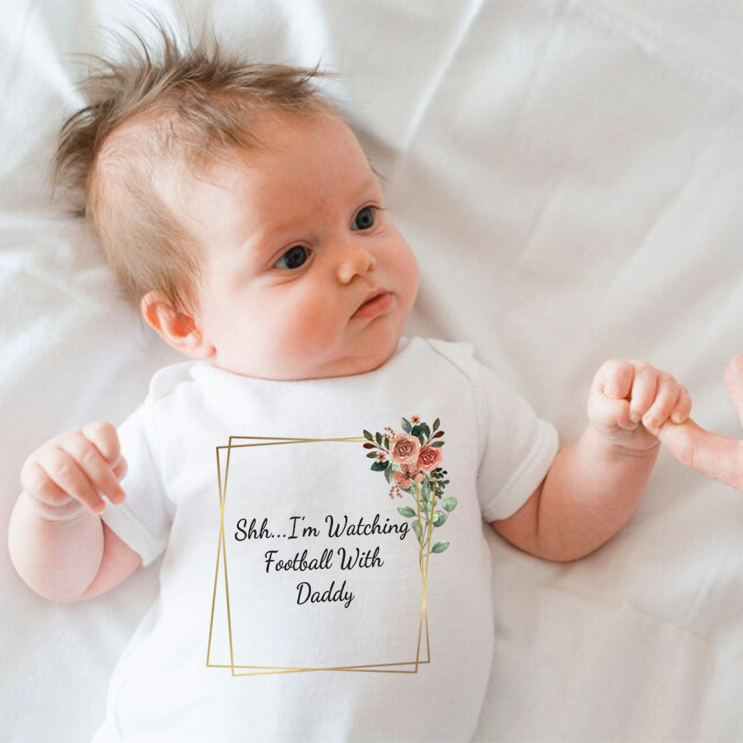 Baby lying on a white blanket wearing a white vest with floral decoration and the phrase "Shh… I'm Watching Football With Daddy" in a geometric frame.